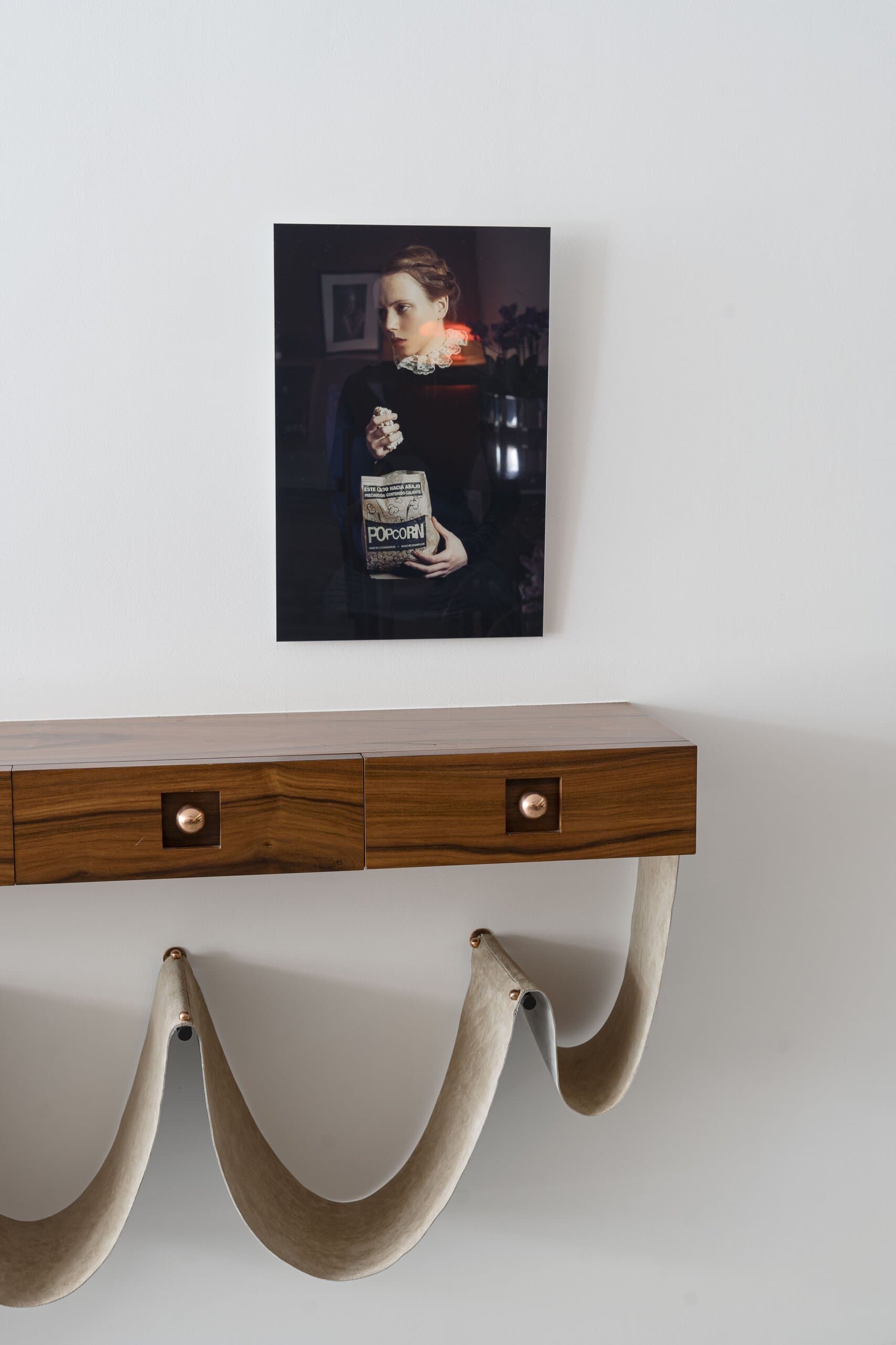 A small timber console with brass pulls hung on a white wall from curved leather straps, a portrait photograph above.