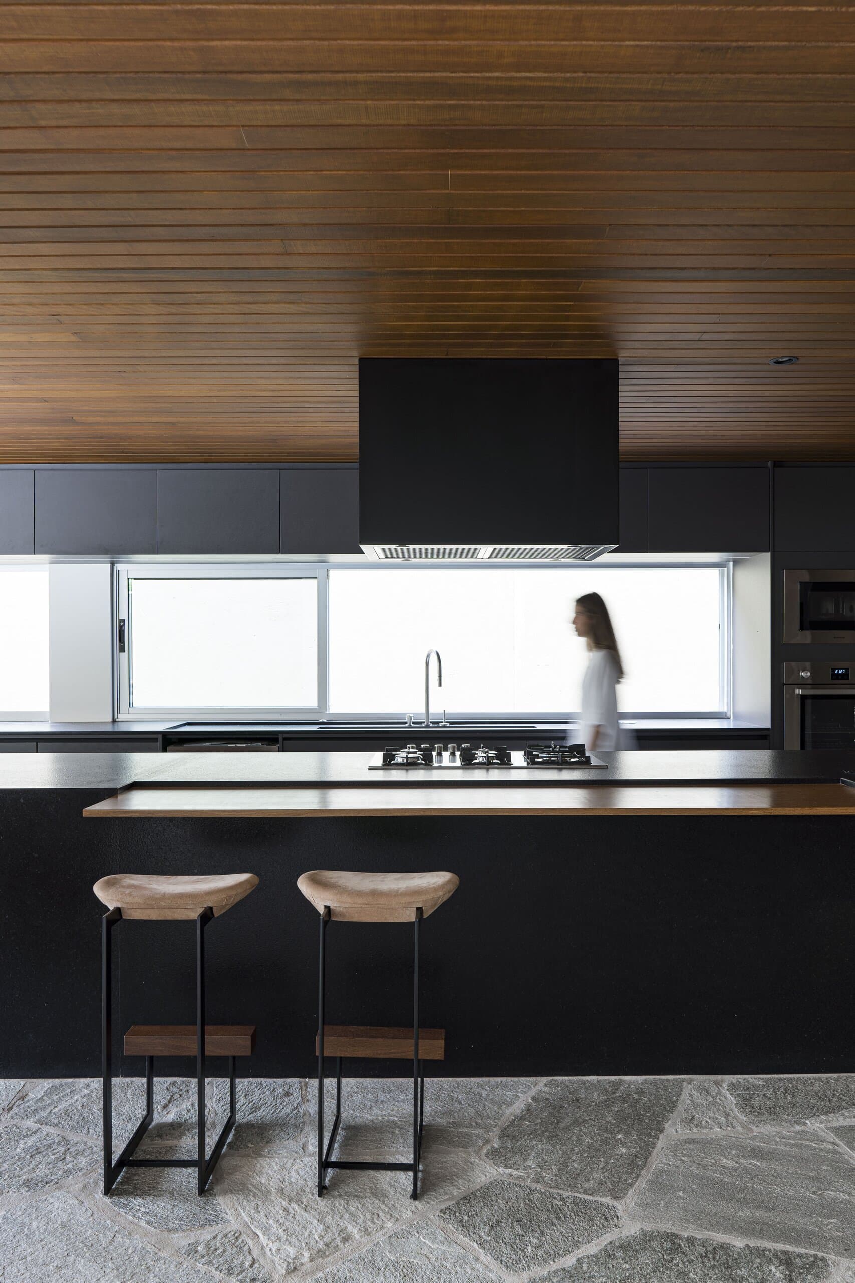 A black kitchen island with two timber stools beneath a slatted wood ceiling, a woman in white passing the window behind.