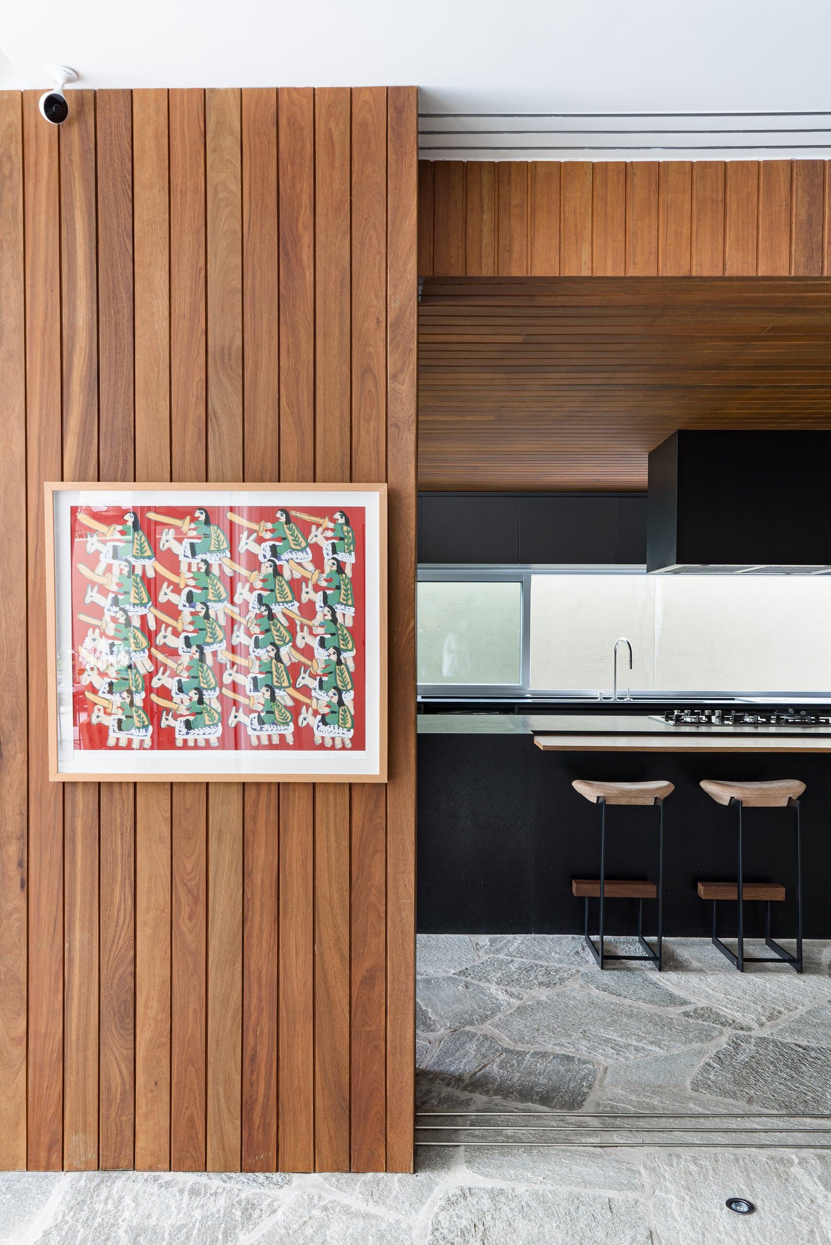 A framed red-and-green print on a vertical timber panel, the black kitchen and stone floor beyond.