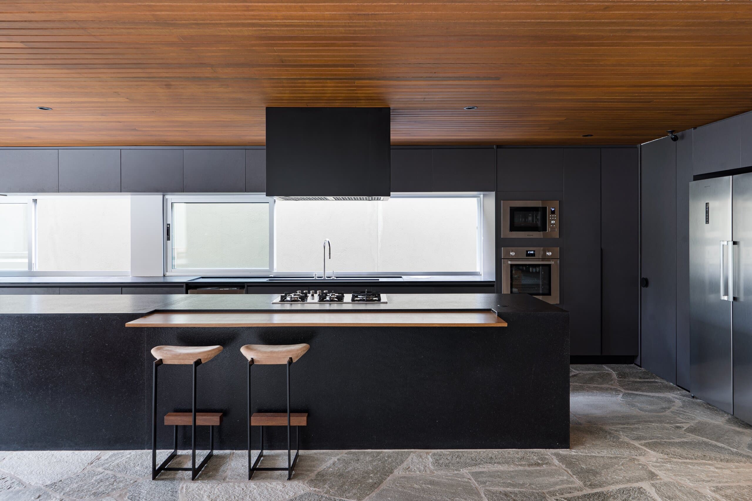 The kitchen head-on: black cabinetry and island, a square black hood and a ribbon window under the timber ceiling.
