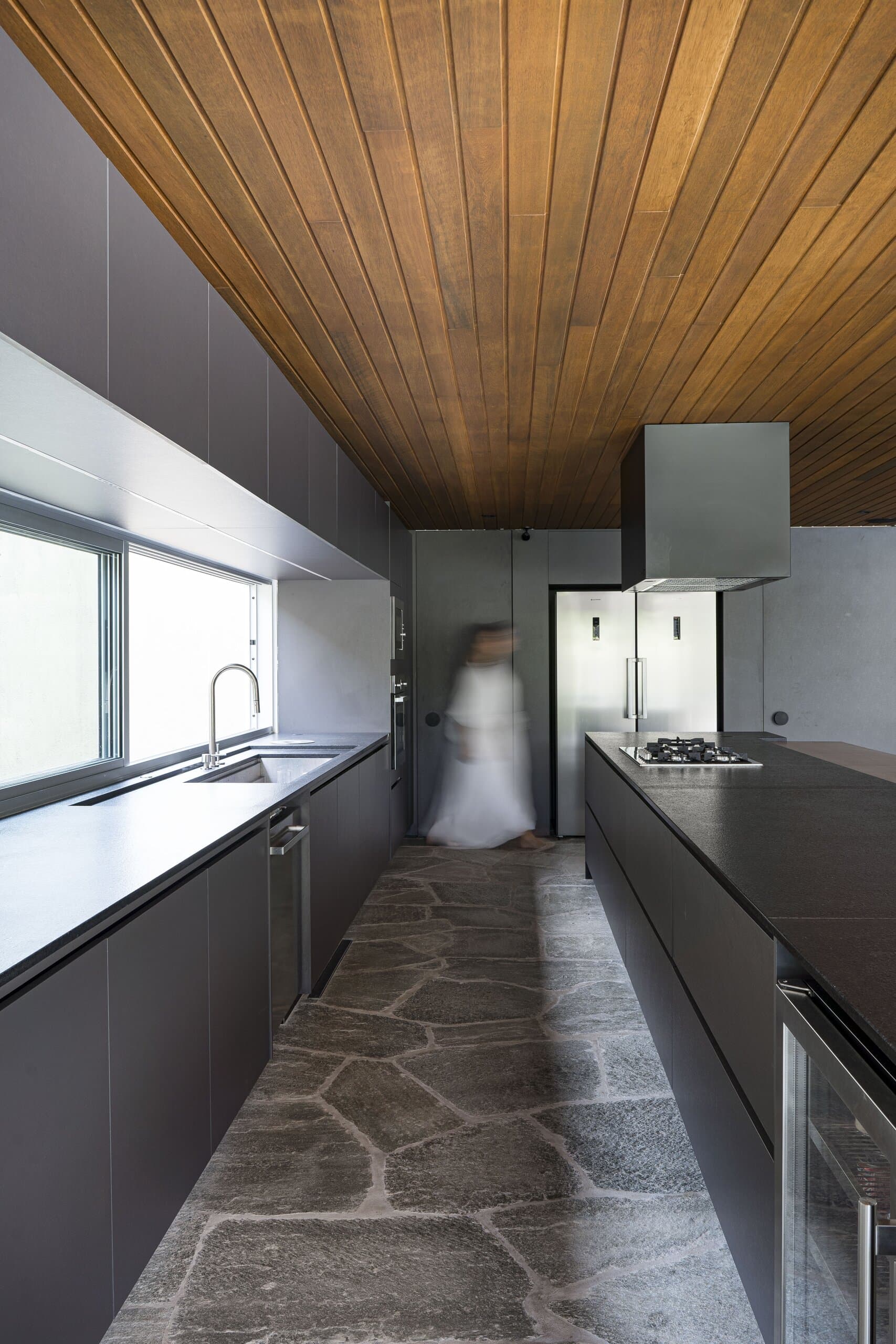 A galley of black cabinets along a window, a figure in white blurred in motion on the irregular stone floor.
