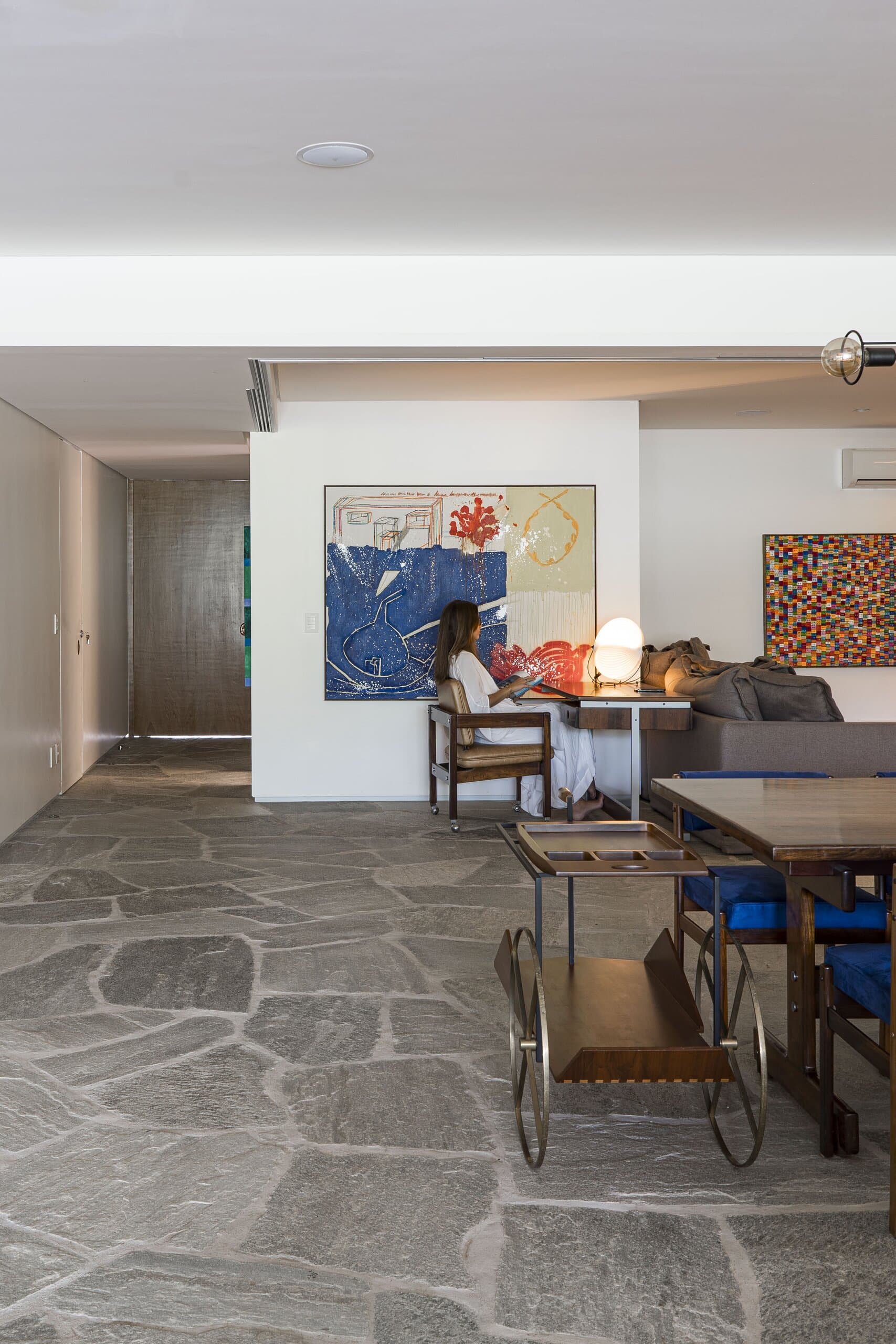 Across the stone floor to the dining area: a woman at a lamp-lit table, a brass bar cart and abstract paintings on white walls.