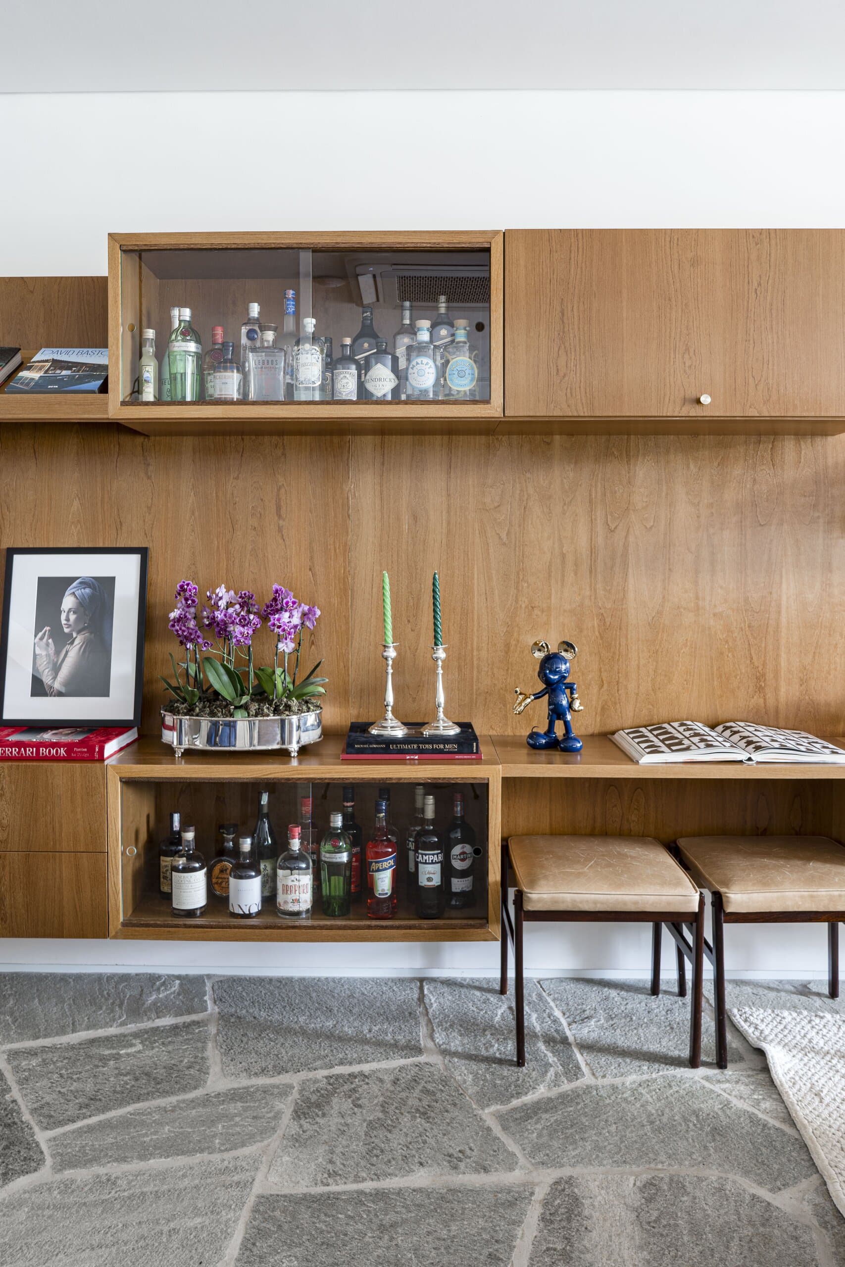 A timber bar unit with a glass cabinet of bottles, a black-and-white portrait, purple orchids and two leather stools.