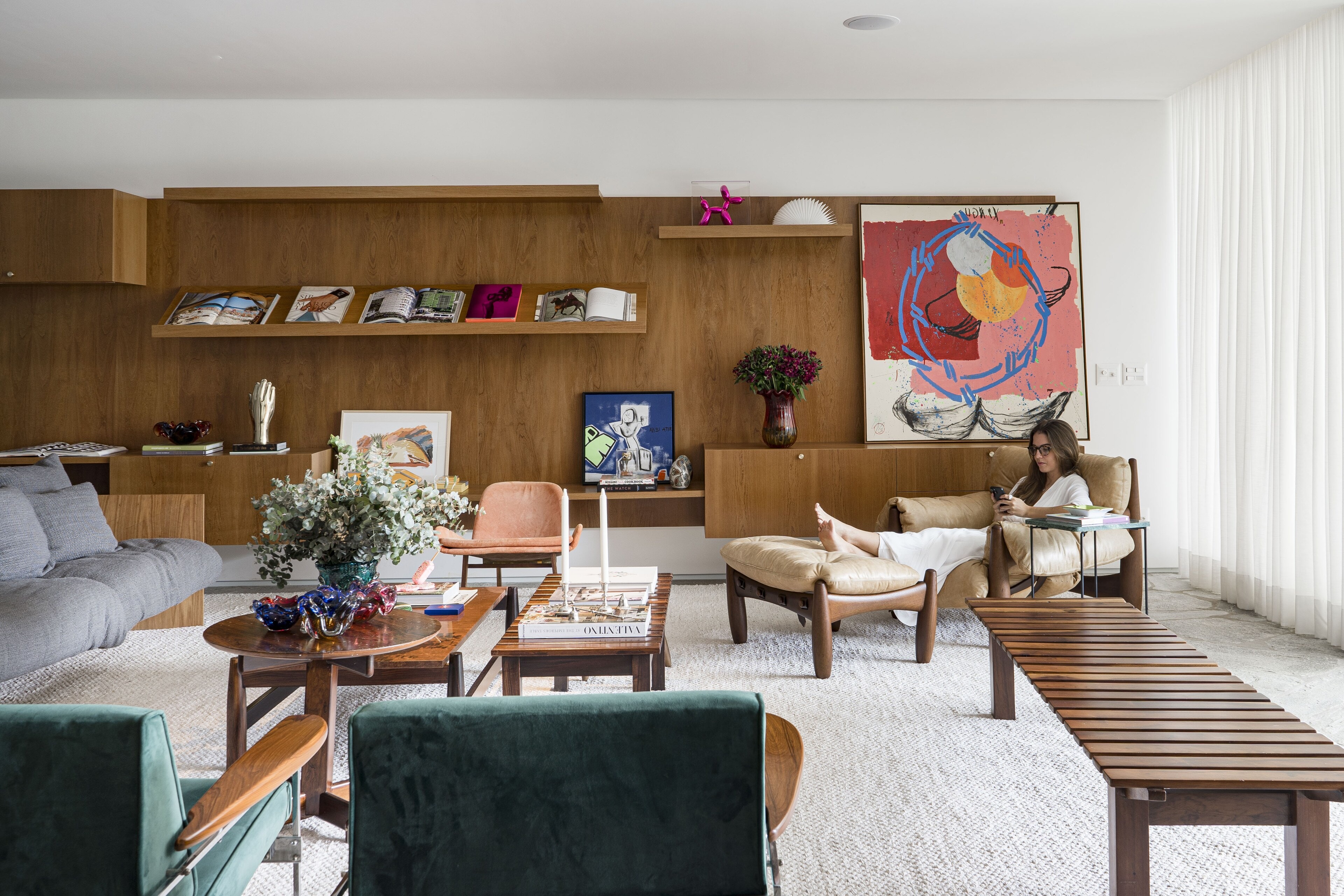 A long living room with a timber wall of shelves and a large red-and-blue painting, a woman reading in a leather armchair beside green velvet chairs.