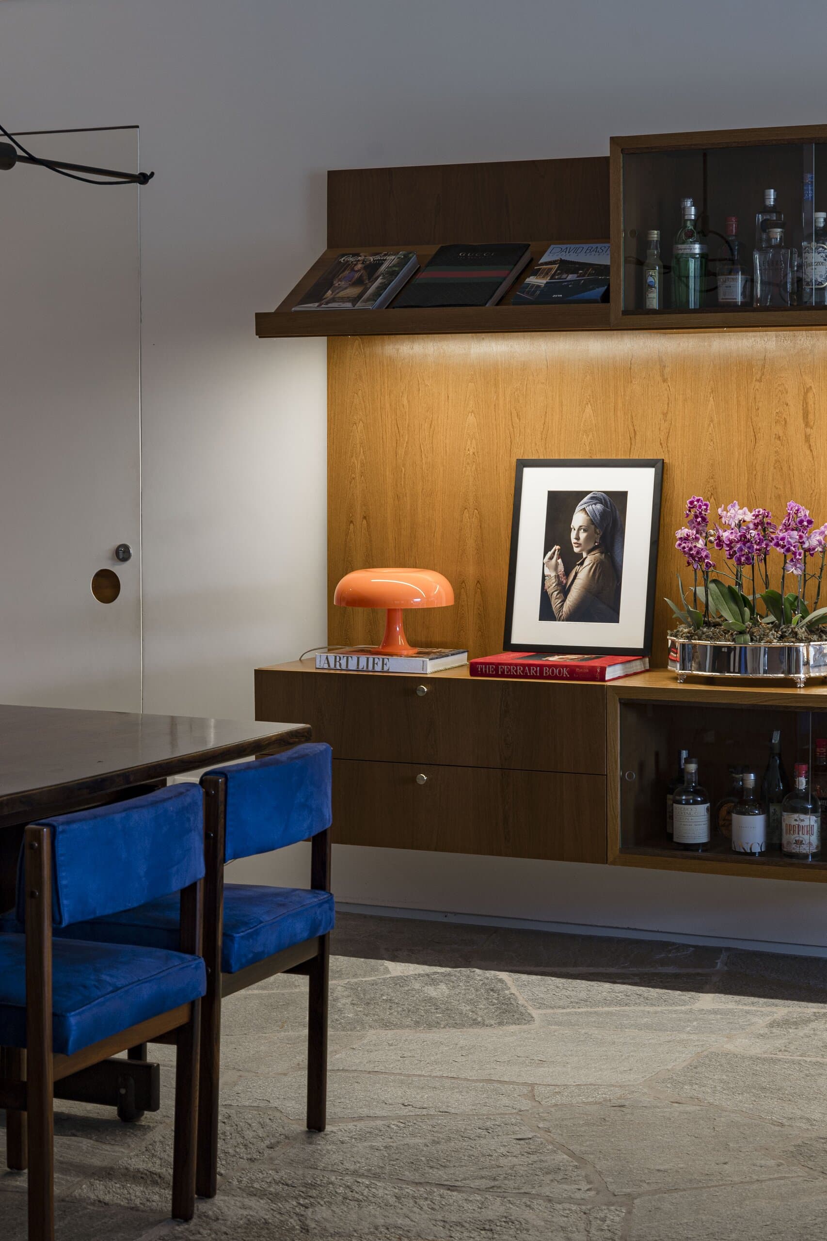 Blue velvet dining chairs below the timber shelves, an orange lamp, the portrait and purple orchids in soft light.