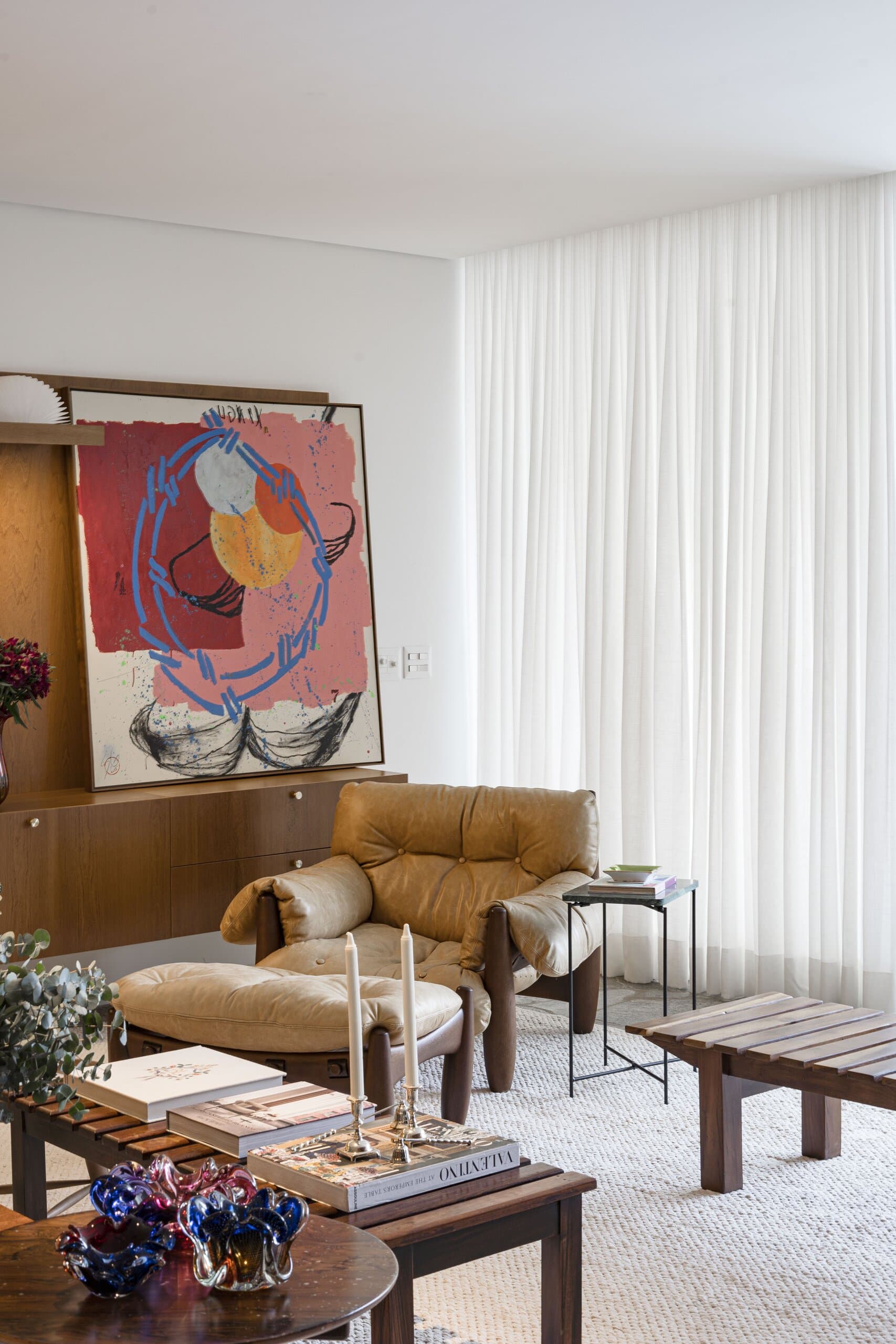 A leather armchair and footstool by tall white curtains, the red-and-blue painting above and books stacked on a timber table.
