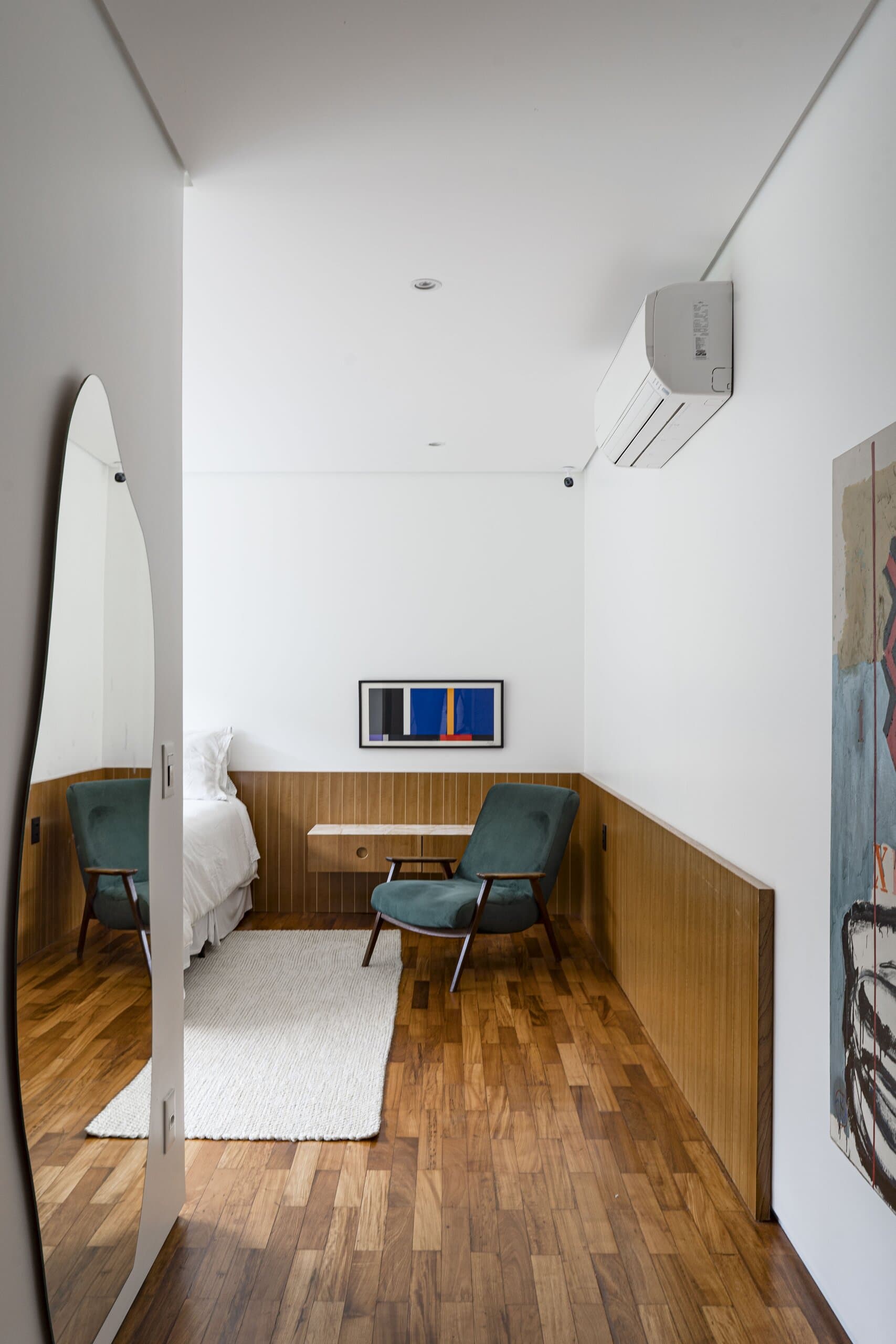 A bedroom with a timber floor and wainscot, a green armchair and a white rug, reflected in a tall arched mirror.