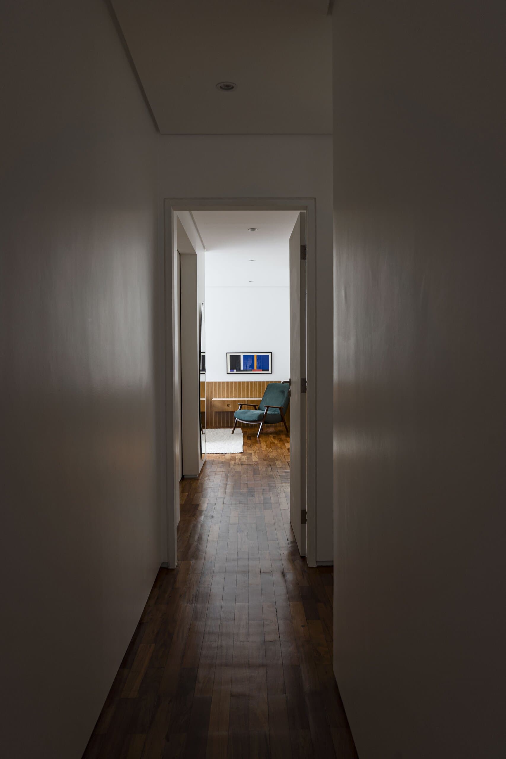 A dark corridor with a timber floor leading to a lit bedroom with a green armchair at the far end.