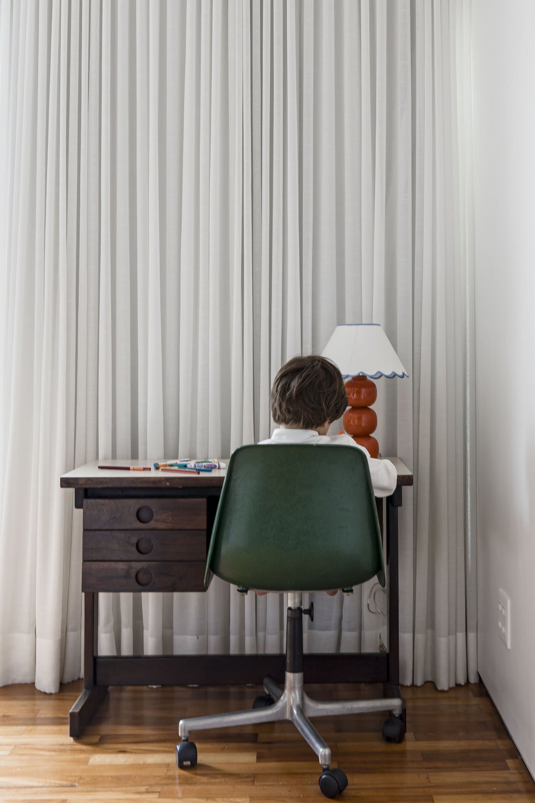 A child at a timber desk in a green office chair, a lamp with a scalloped shade and white curtains behind.