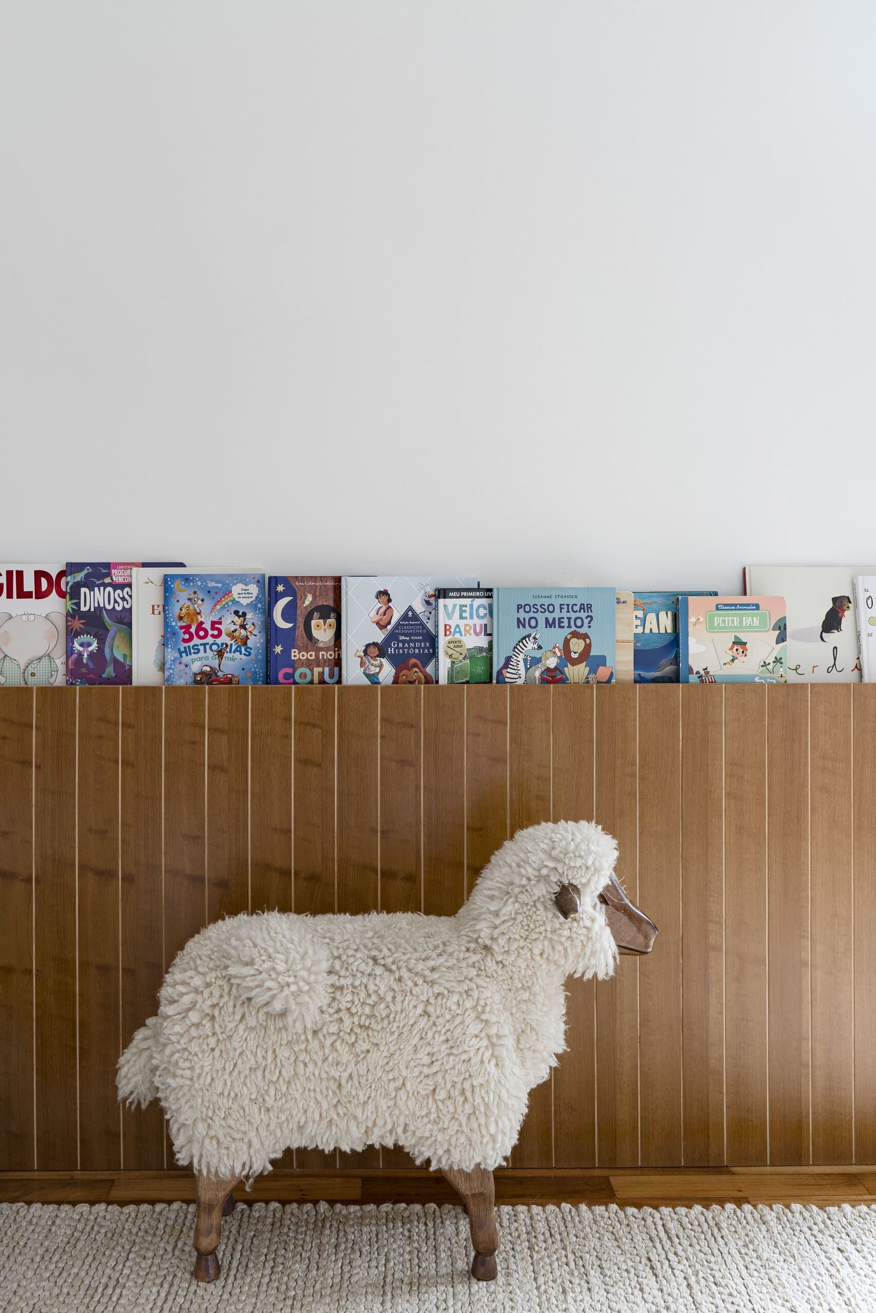 A woolly toy sheep on a cream rug before timber panelling, a row of children's books on the ledge above.