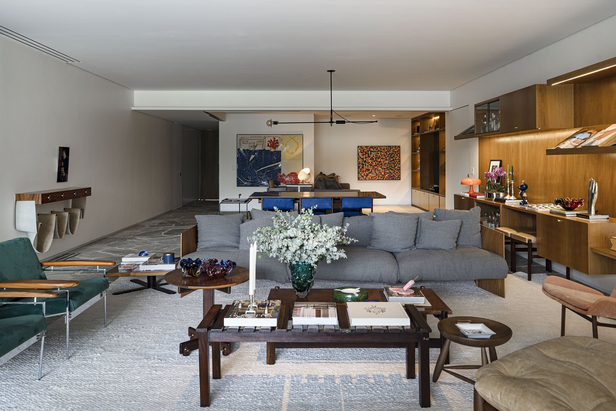 The full length of the living room: grey sofa, dark timber coffee table, green armchairs and a white bouquet, the dining table and shelves beyond.