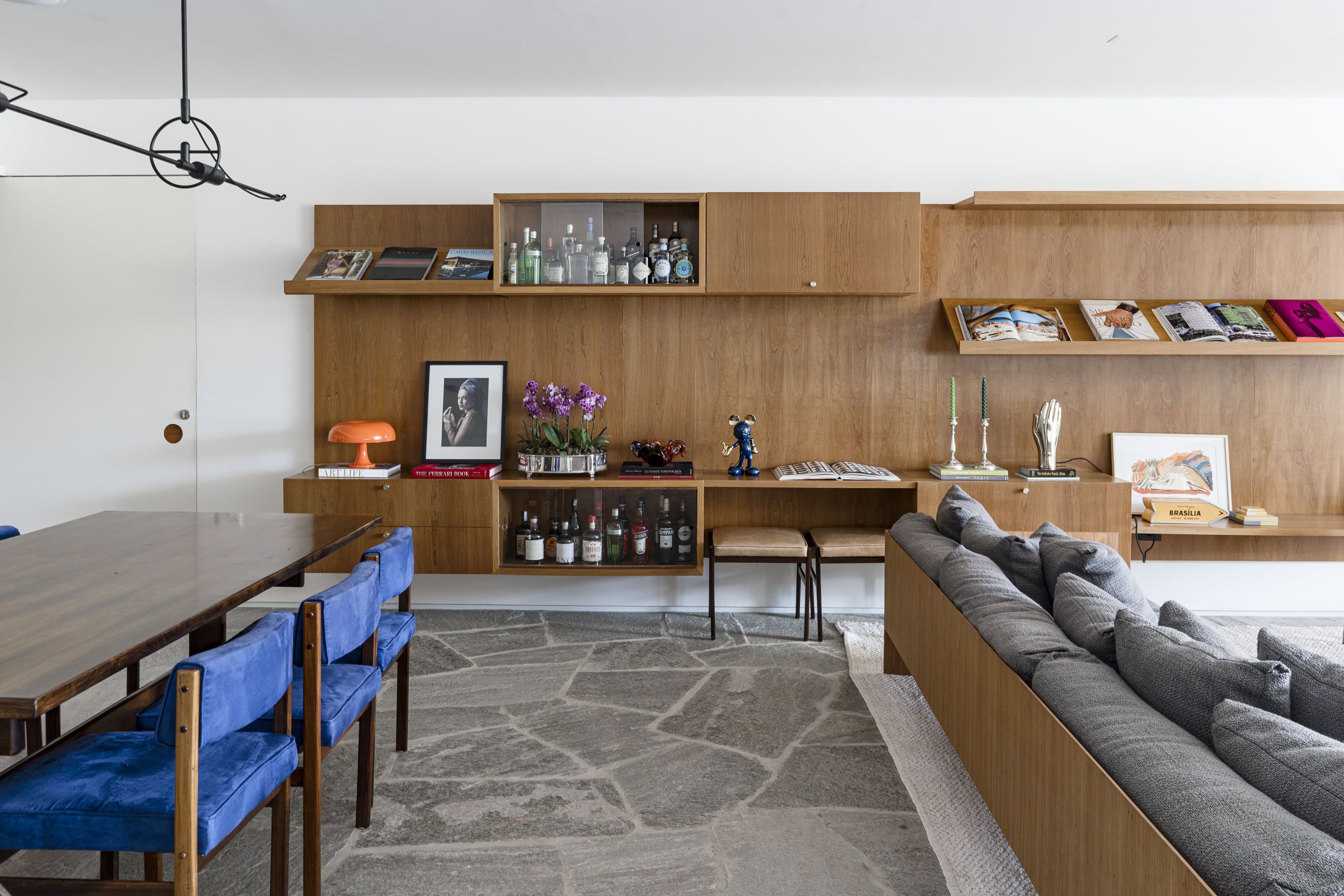 A long living room: grey sofas and blue velvet dining chairs beneath a timber wall of shelves, books and bottles, on crazy-paved stone.