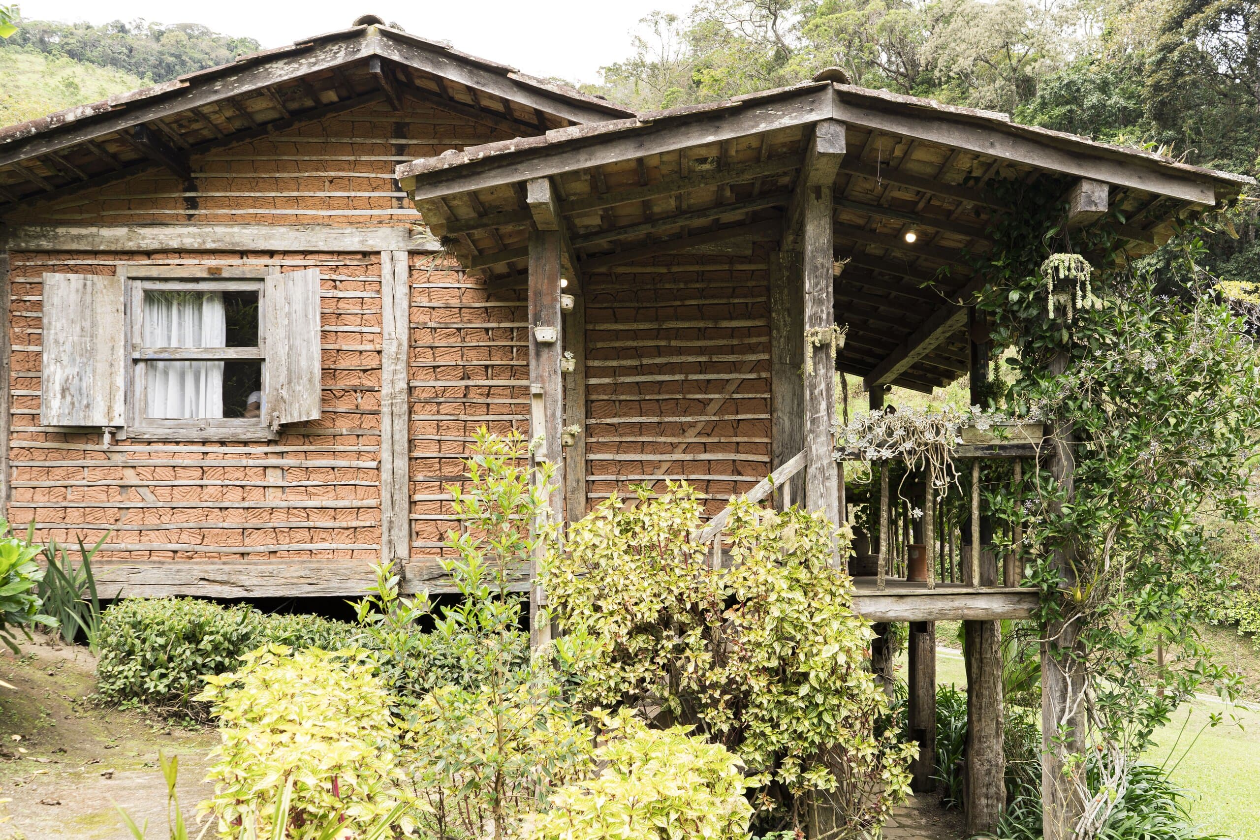 A weathered timber chalet on stilts with a deep porch, wrapped in garden planting and hillside trees.
