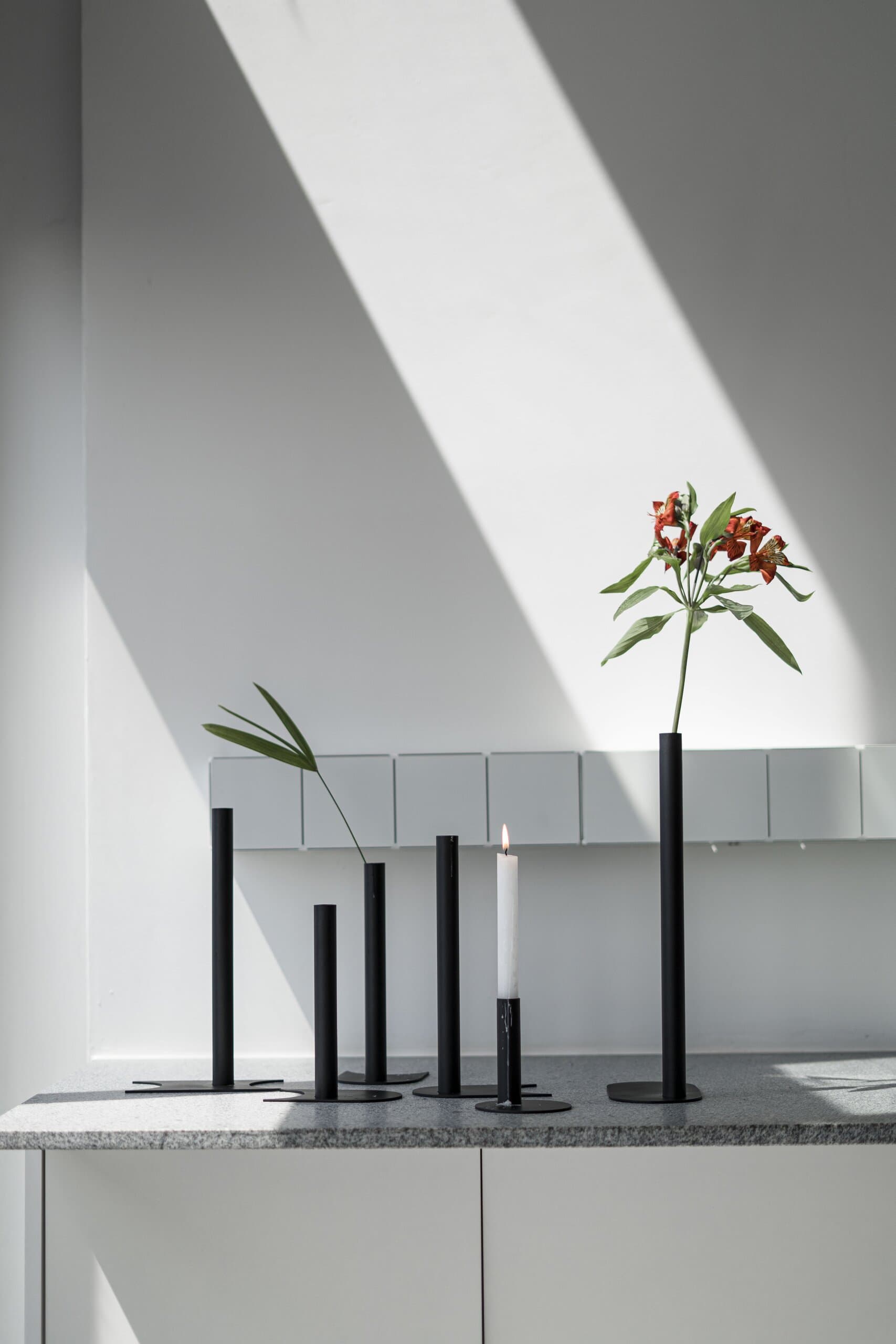 Slender black vases holding single red lilies on a white console, cut by a diagonal band of sunlight.