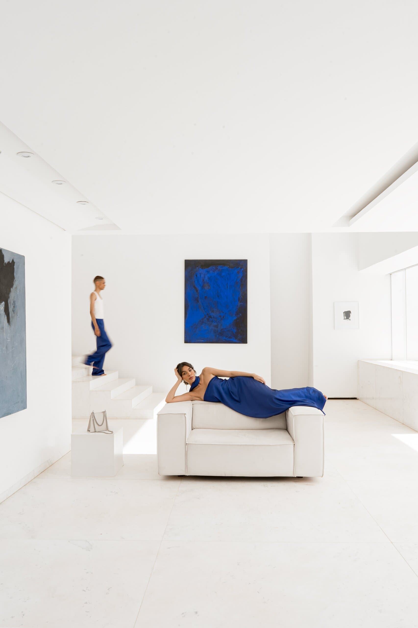 A white gallery-like room: a woman in blue lies across a white armchair while another walks down the stairs, a blue painting on the wall.