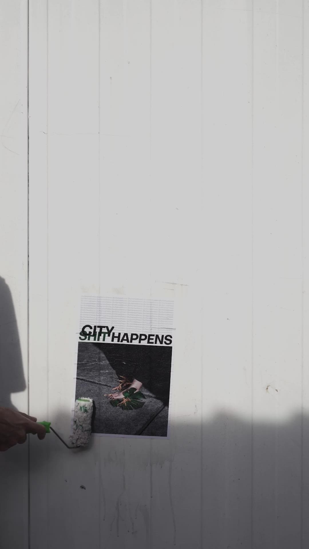A "City happens" poster is pasted onto a white boarded wall with a paint roller, half in shadow.