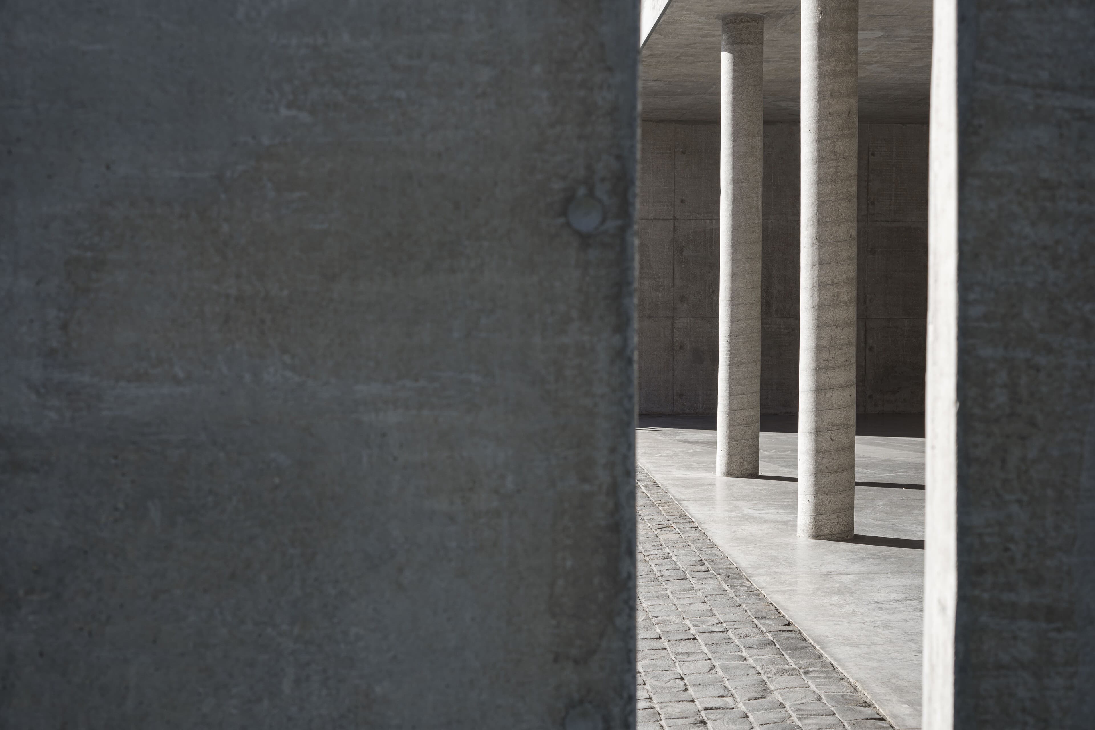 Through a gap in a concrete wall: slender round columns, a polished floor and a strip of cobbles in raking light.