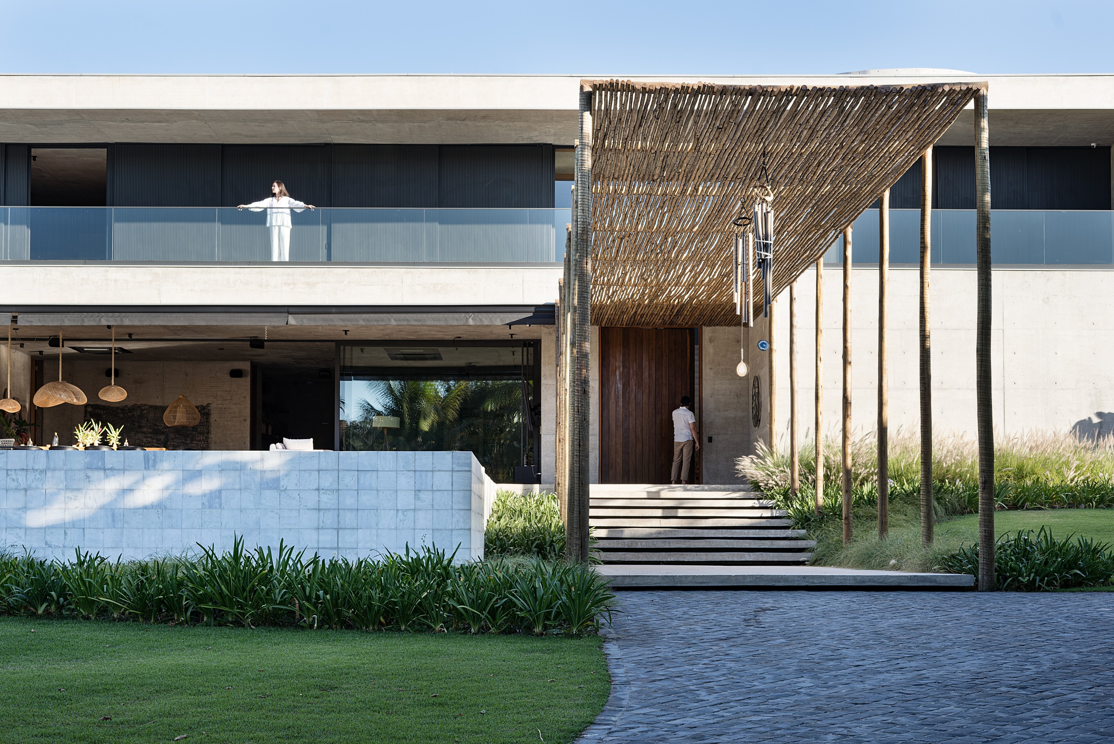 SJ House: a long concrete house beneath a flat roof, a woman at the glass balustrade above and a man at the timber door under a pergola of branches.