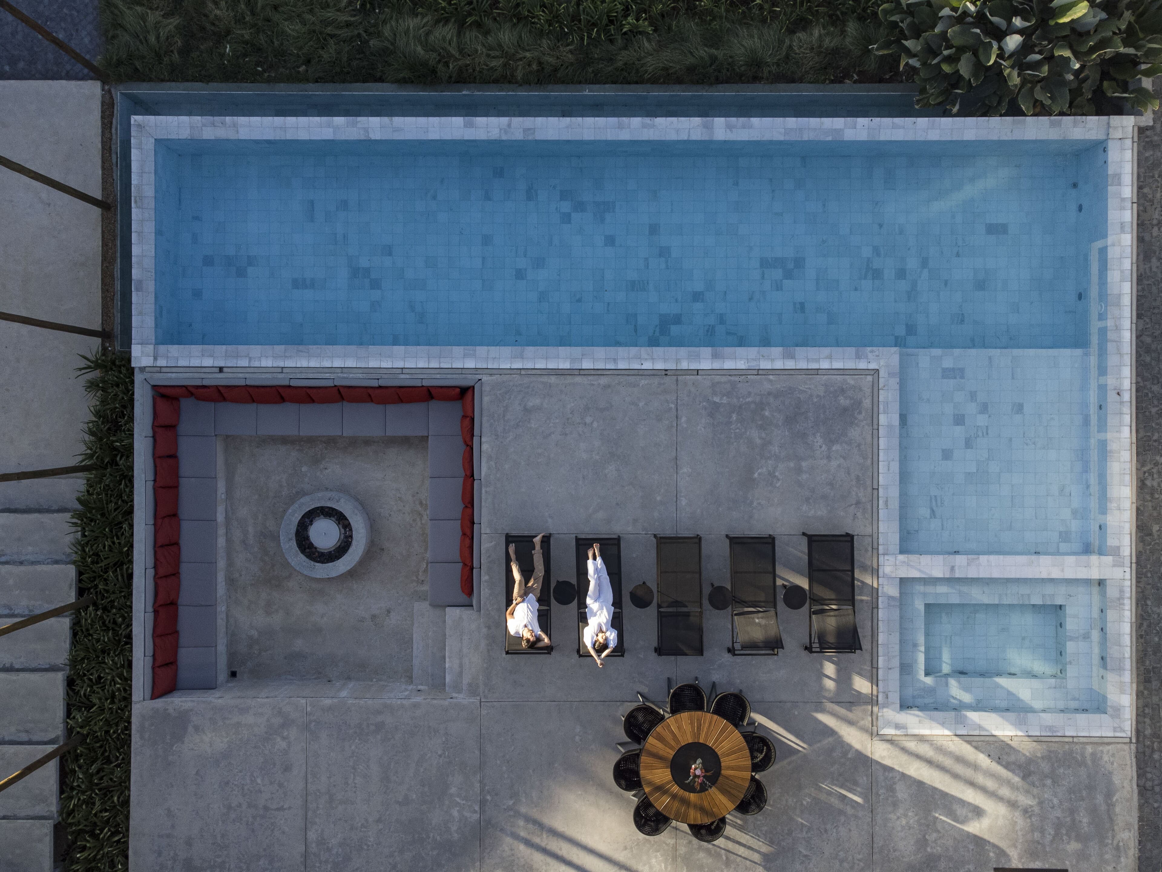 From above: the long pool and its terrace — a sunken fire pit lined with orange cushions, loungers, a round timber table and two figures.