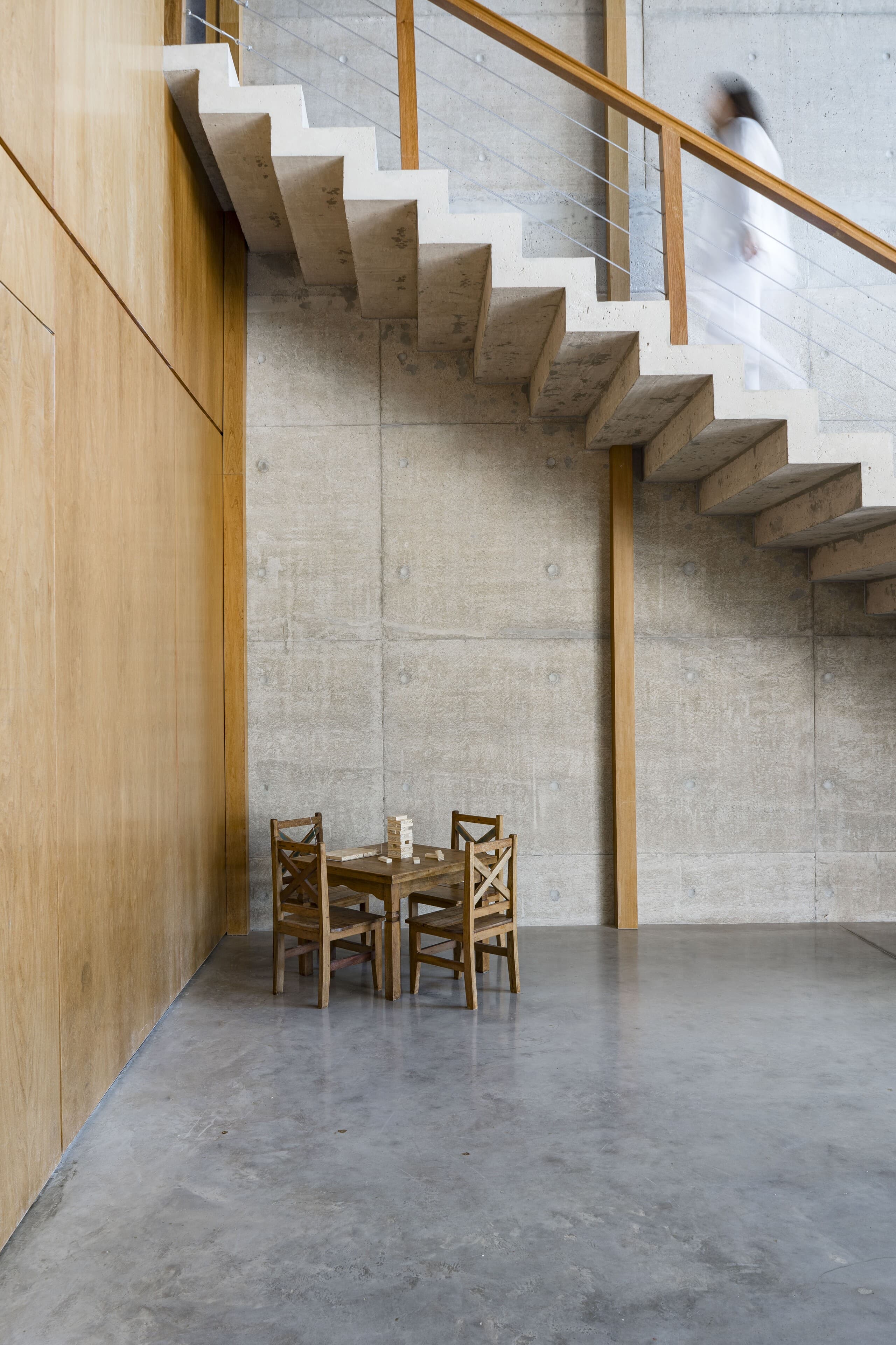 A concrete stair rises beside a timber wall, a small table and chairs below, a figure blurred in motion.