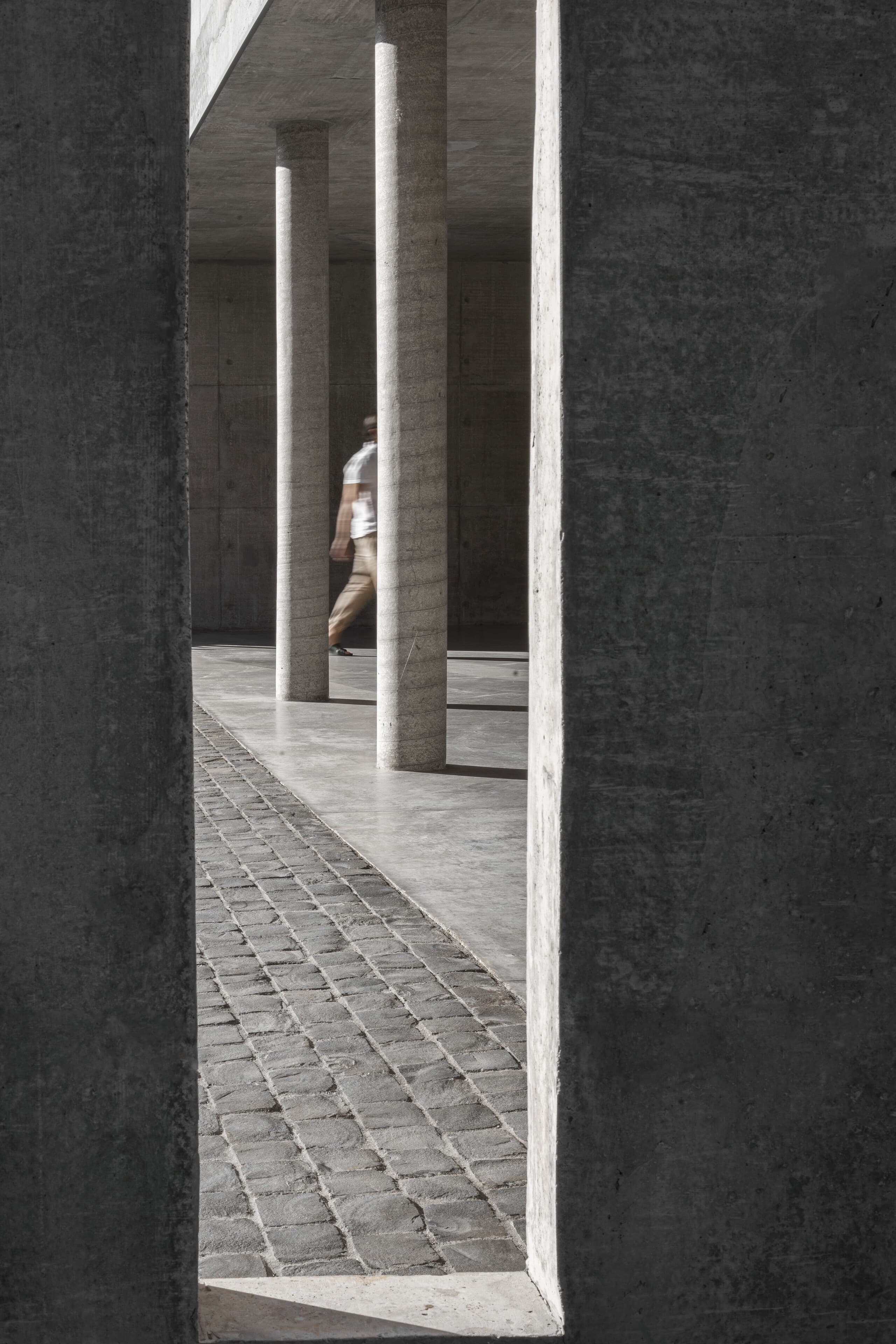 Between two concrete walls: a colonnade of round columns and a cobbled path, a figure passing.