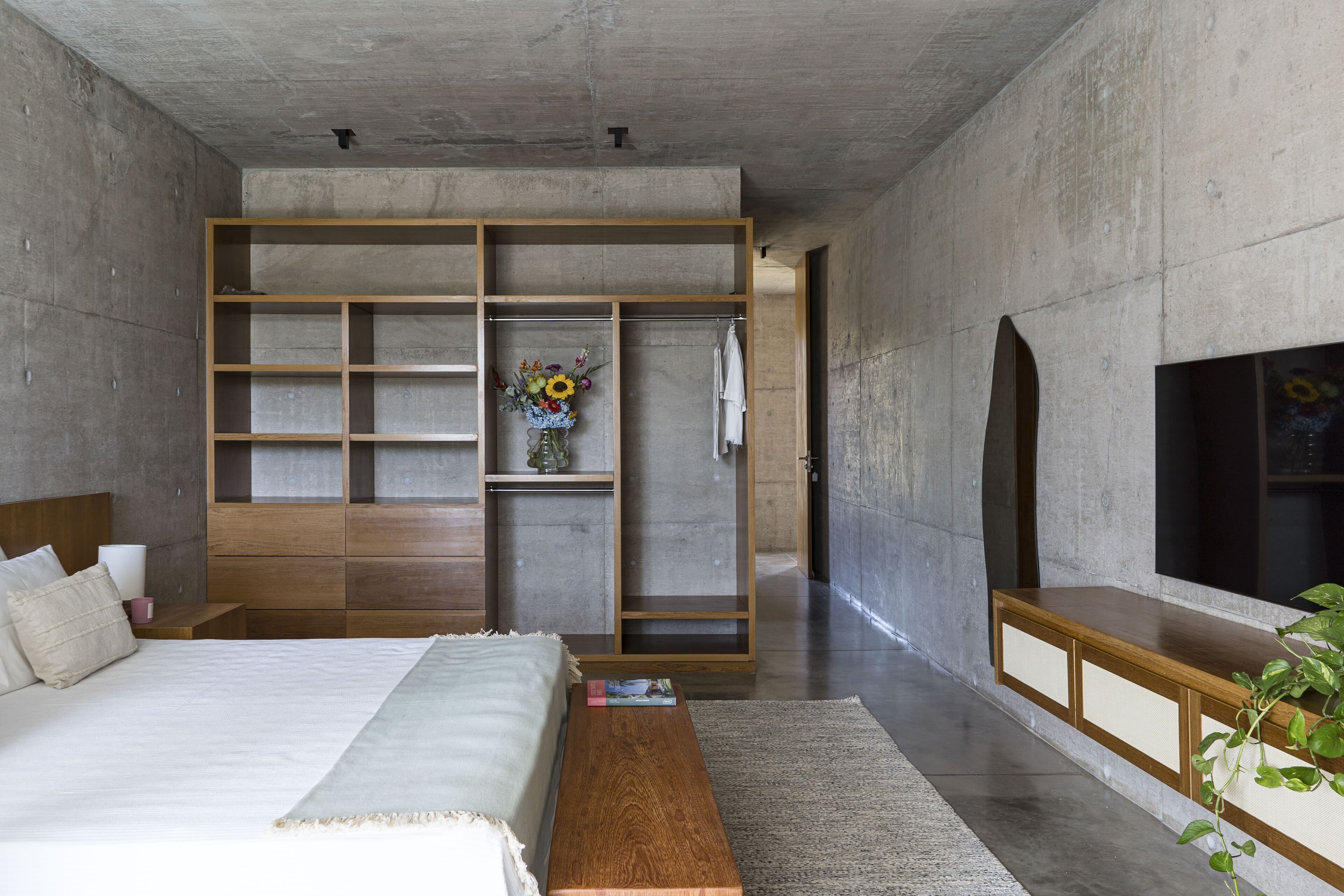 A bedroom in board-formed concrete with timber shelving, a low bench, a woven rug and flowers in a glass vase.