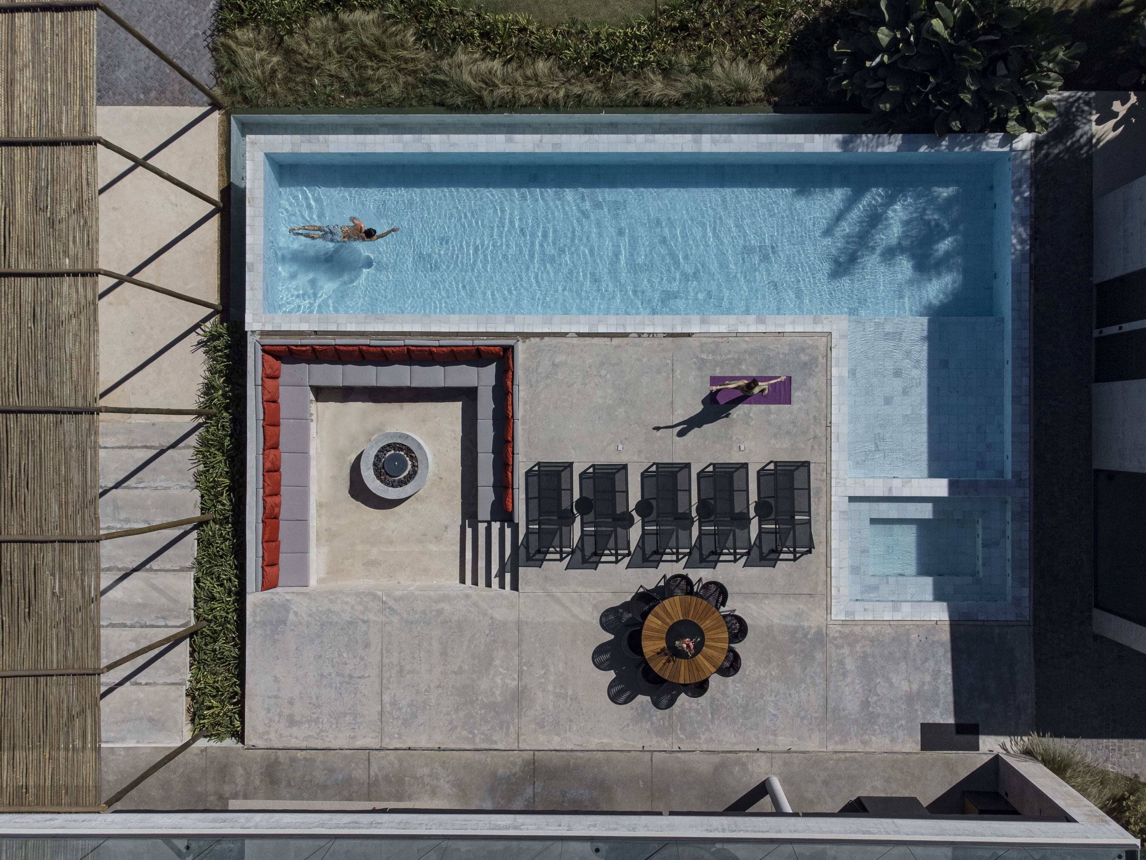Vista de cima: uma pessoa nadando na longa piscina azul, uma mulher deitada num tapete roxo no terraço de concreto, espreguiçadeiras, uma mesa redonda de madeira e o fogo de chão rebaixado com borda vermelha.