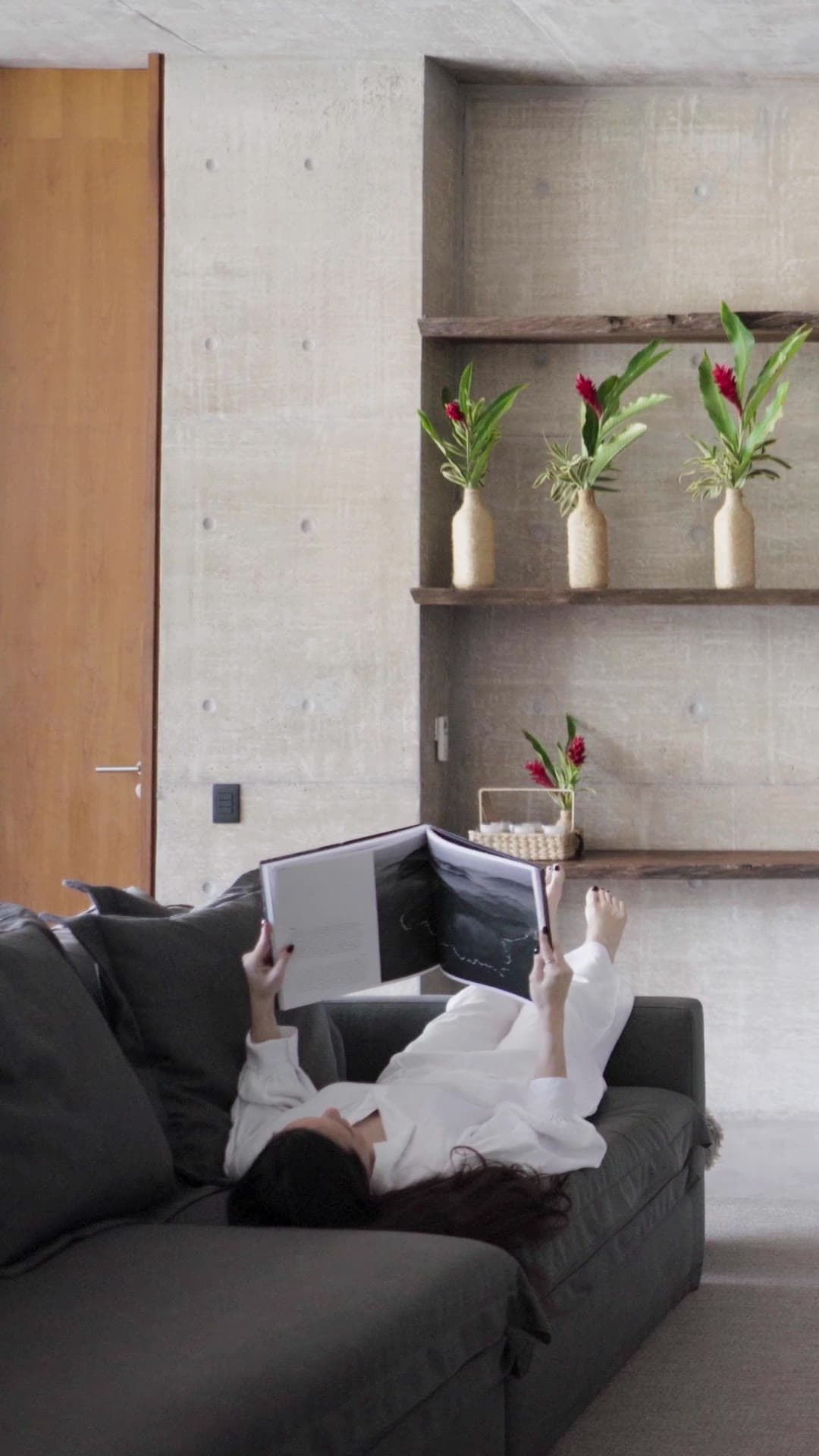 SJ House — a second vertical cut: a woman reading on a grey sofa, the pergola path, broad-leaved planting and a long concrete corridor.