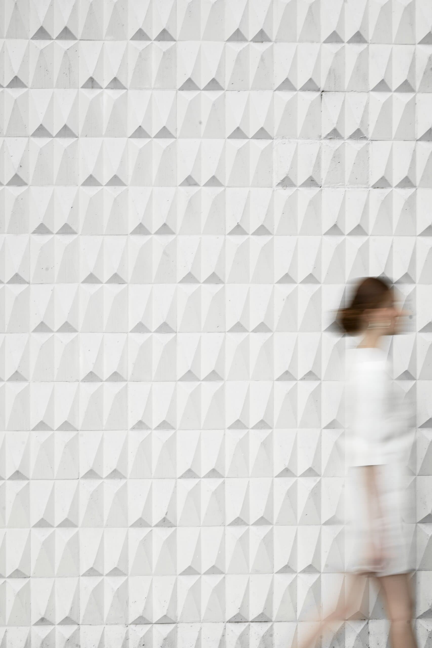 Uma mulher de branco passa desfocada diante de uma parede de azulejos brancos facetados.