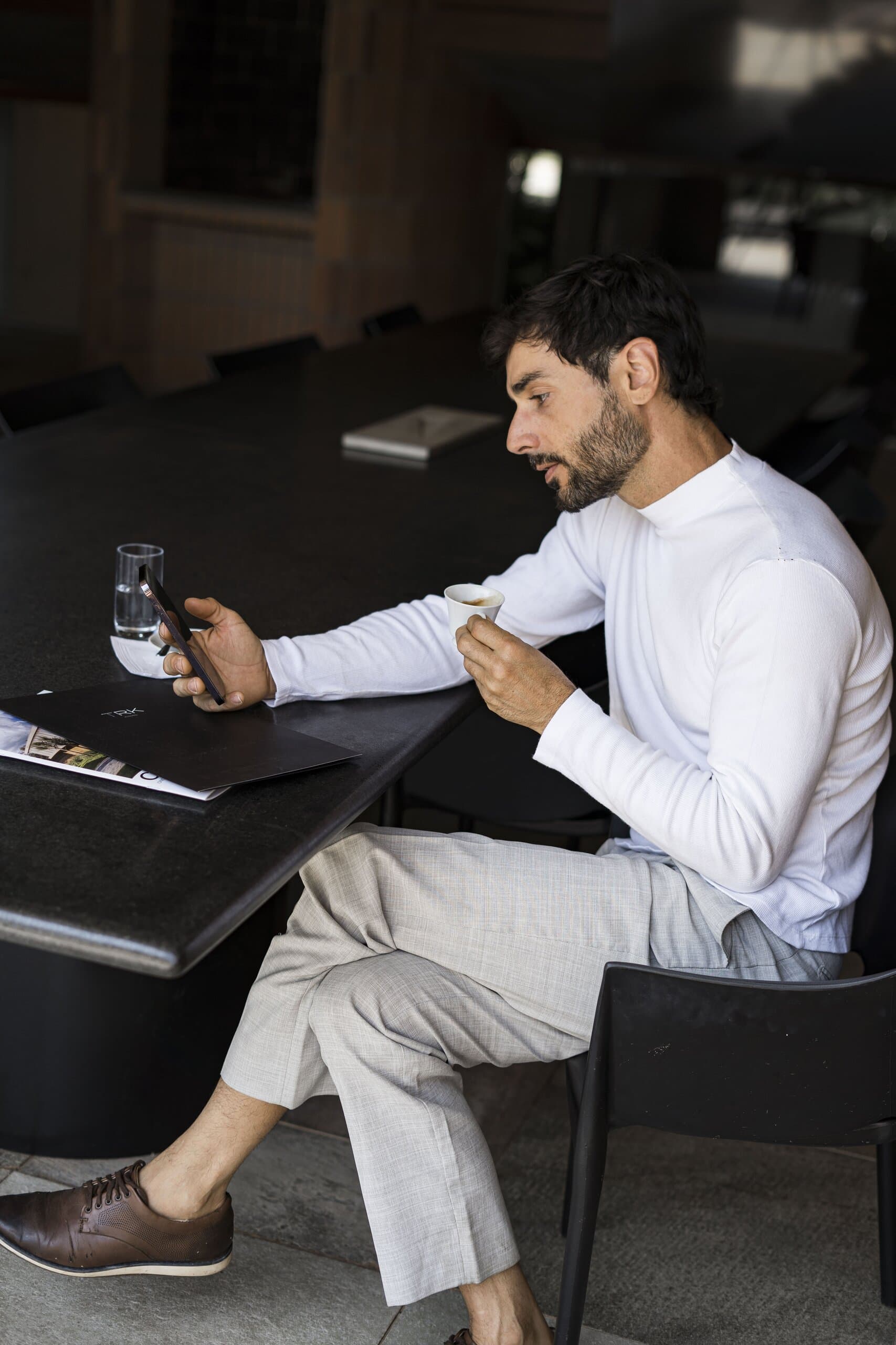 A bearded man in a white roll-neck sits at a dark table with a phone and a glass of water, an open magazine beside him.