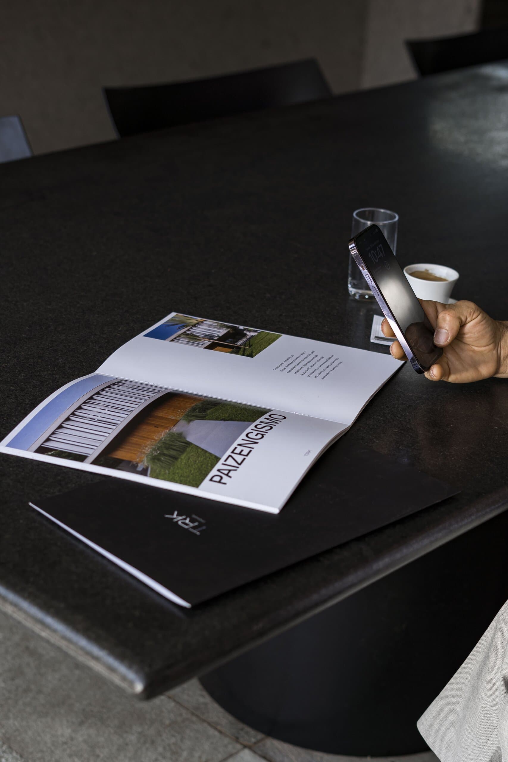 Uma revista aberta e uma pasta preta sobre a mesa escura, uma mão segurando o celular ao lado de um café.