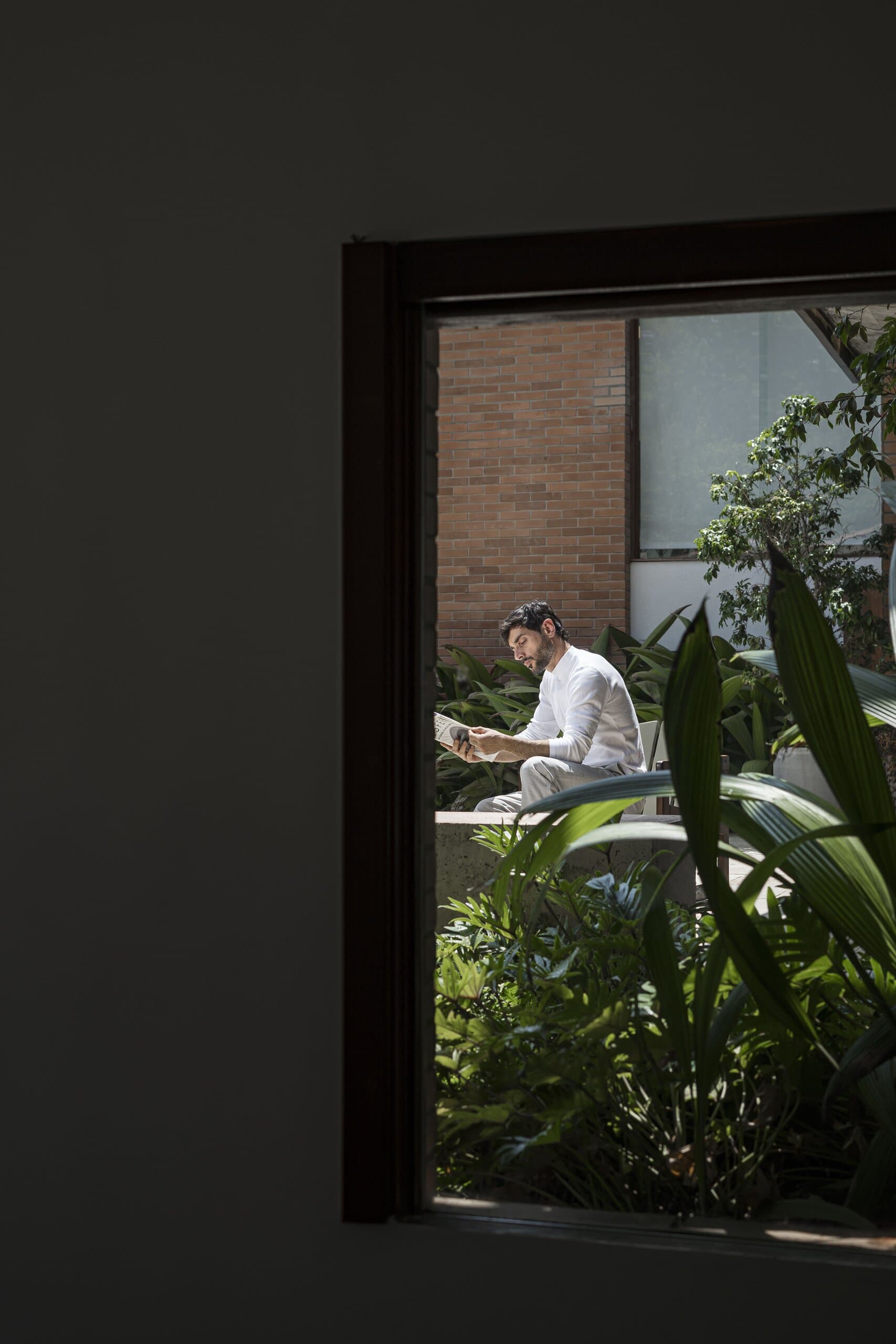Por uma porta escura: um homem de branco lendo num jardim ao lado de um prédio de tijolos.