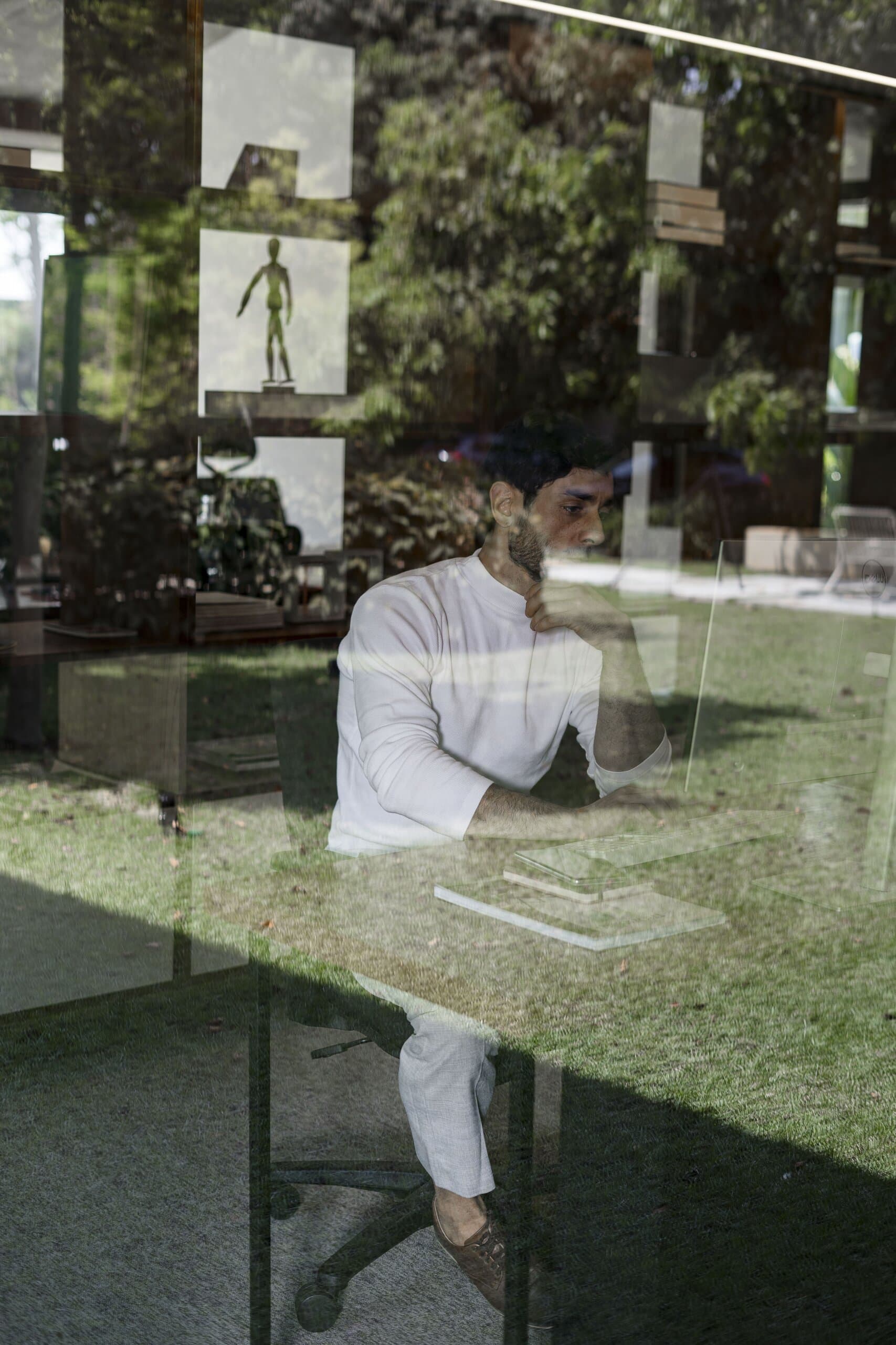 Através do vidro, um homem de branco trabalhando à mesa, com o jardim e quadros refletidos sobre ele.