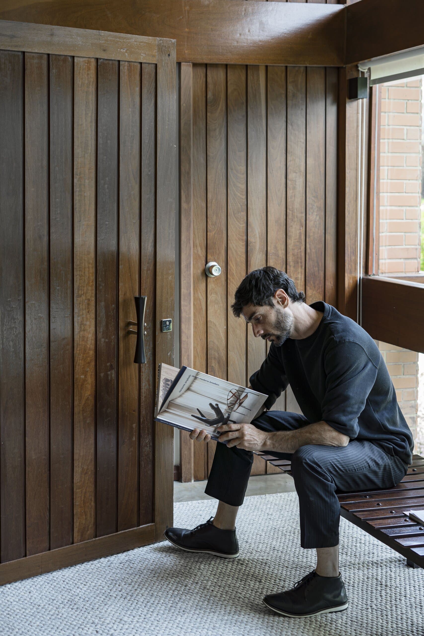Um homem de preto lê uma revista num banco de madeira ao lado de uma porta alta de madeira.
