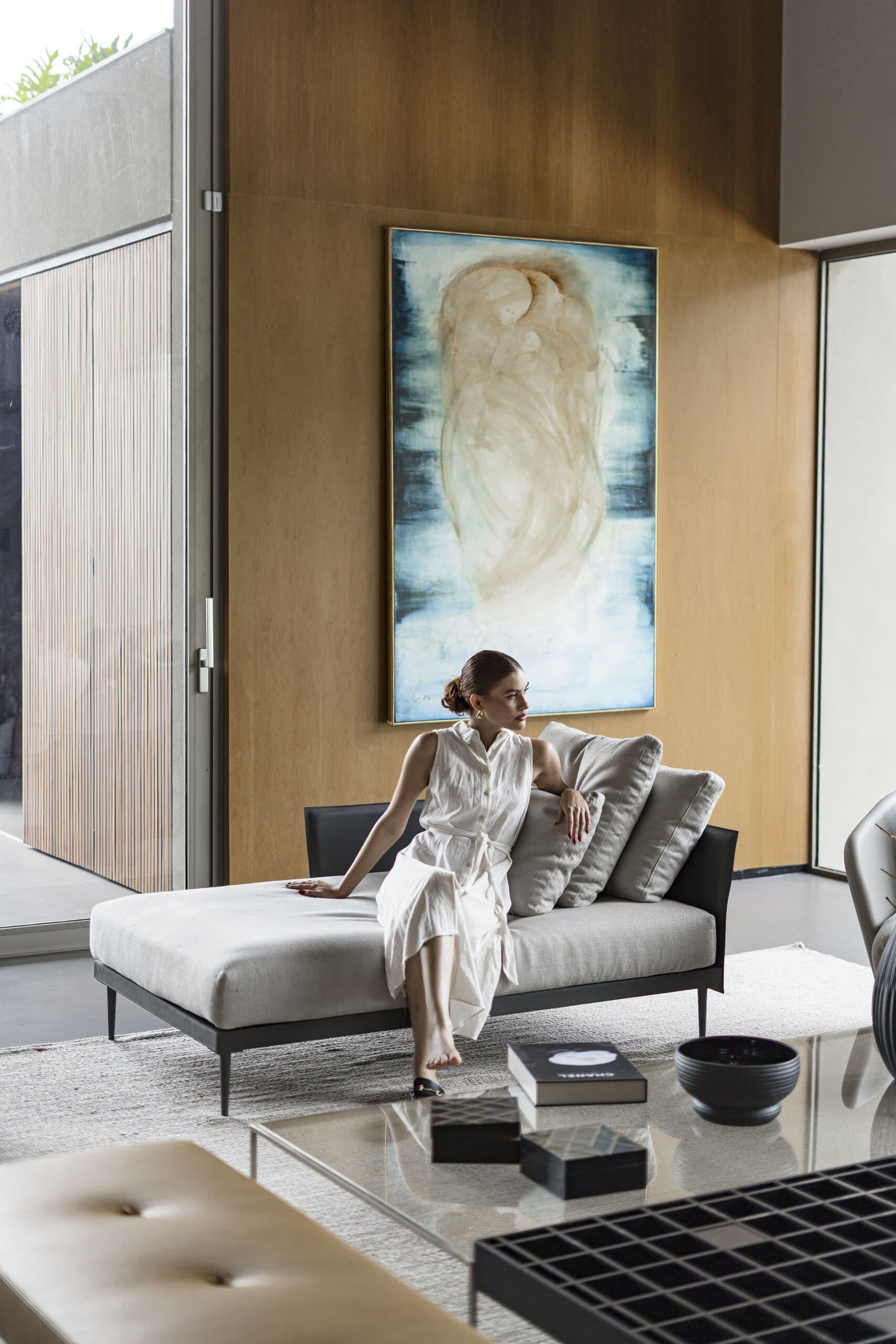 Uma mulher de vestido branco sentada numa chaise cinza sob uma grande pintura numa parede de madeira.