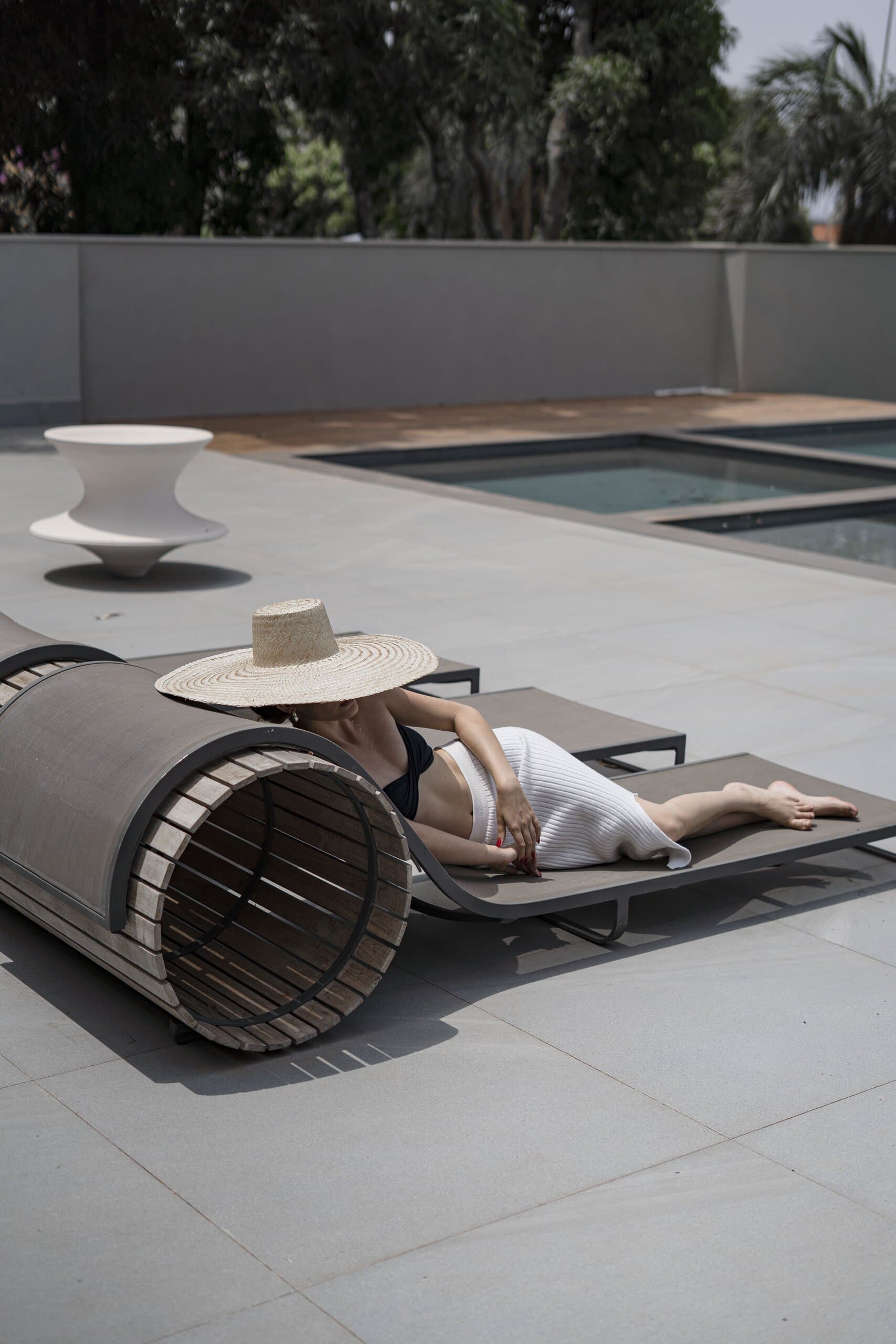 Uma mulher de chapéu de palha deitada numa espreguiçadeira junto à piscina, com piso cinza e árvores ao redor.