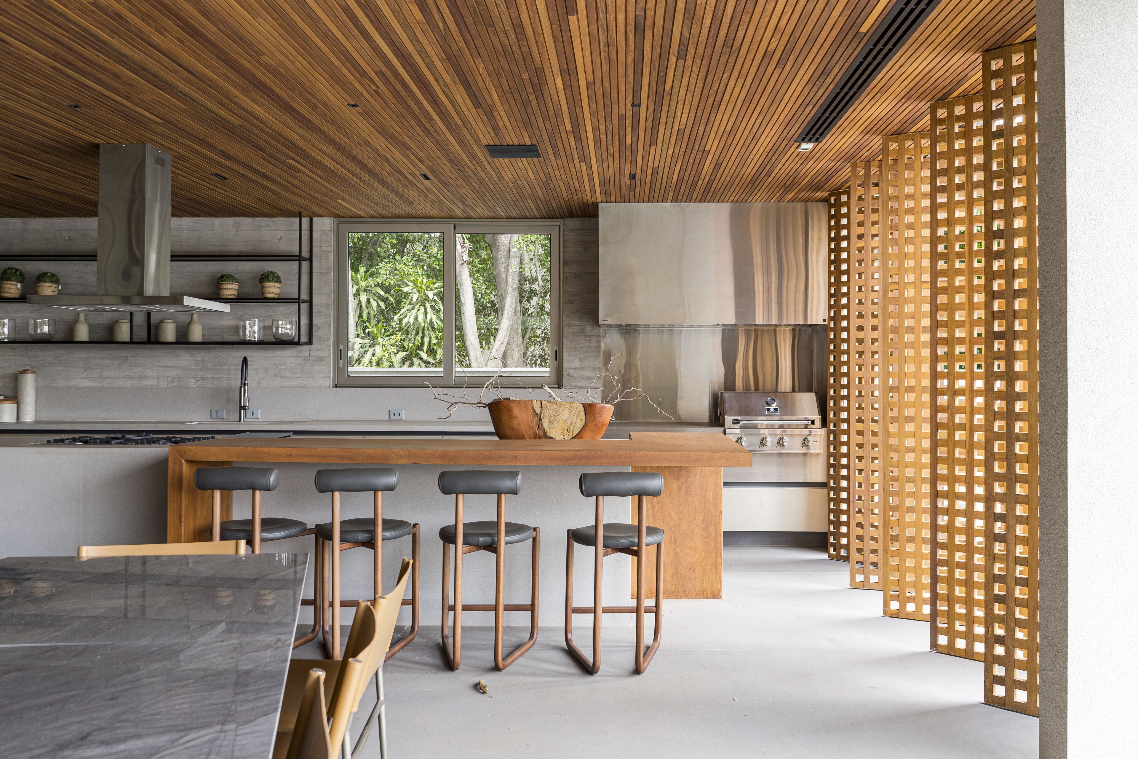 Uma cozinha sob forro de ripas de madeira: ilha de madeira com quatro banquetas, coifa de aço, prateleiras abertas e um painel vazado de madeira à direita.